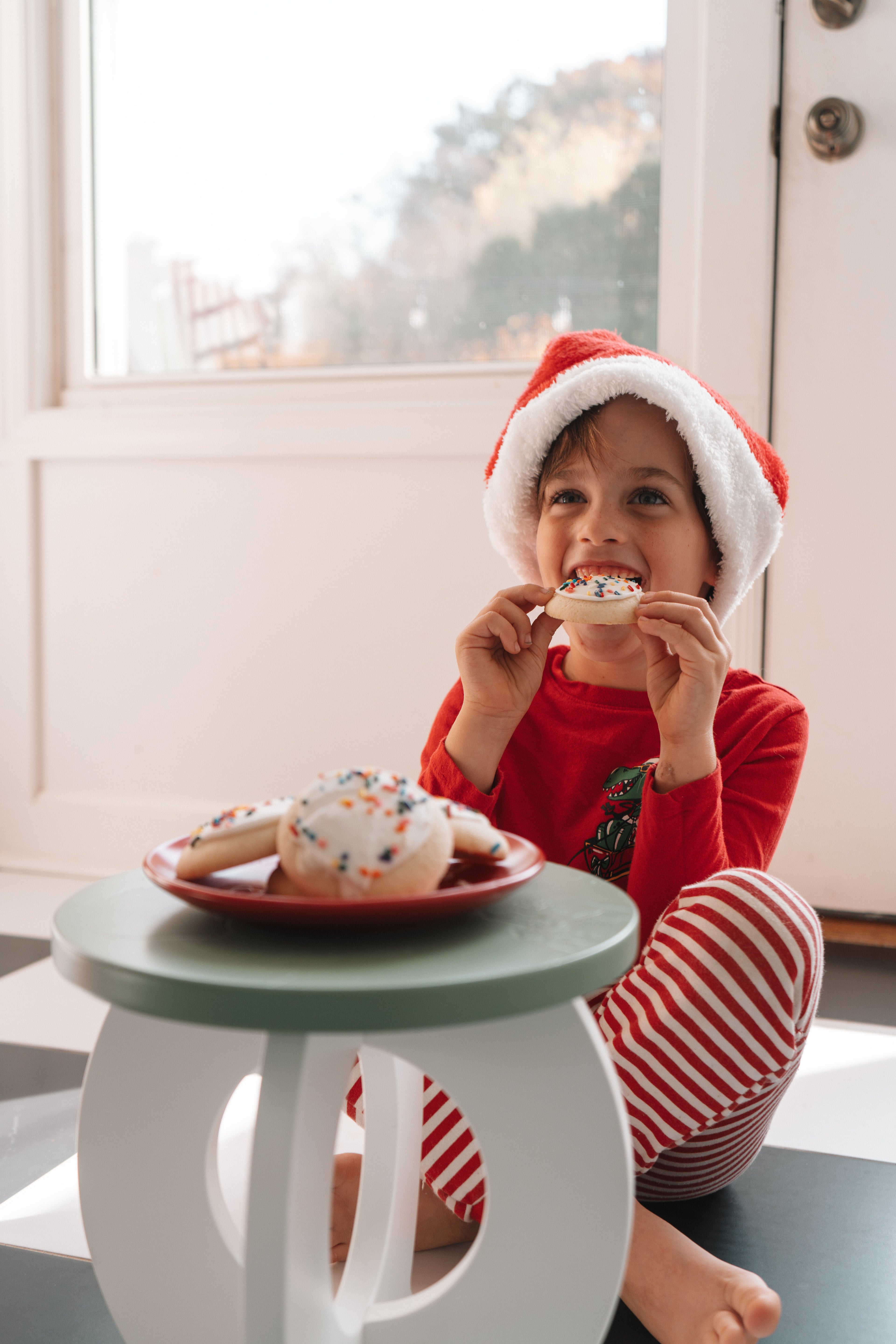 Child in a Santa hat eating a cookie with a plate of cookies and a glass of milk on a small table.